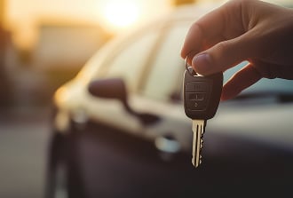 Clé de Voiture à la Main : Démarrage et Liberté Main tenant une clé de voiture moderne avec télécommande, devant un véhicule sombre au coucher du soleil.