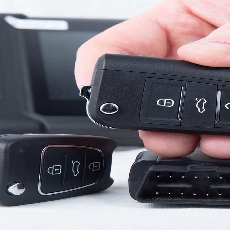 Main tenant une clé de voiture électronique noire de type Volkswagen avec des boutons de verrouillage et de coffre, près d'un autre boîtier de clé et d'un connecteur OBD2.