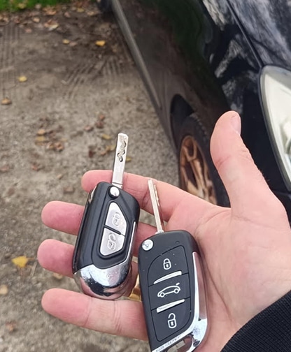 Clés de Voiture Peugeot : Doubles & Remplacement Main tenant deux clés de voiture Peugeot pliantes, l'une chromée et l'autre noire avec boutons verrouillage/déverrouillage et coffre, devant une voiture noire.