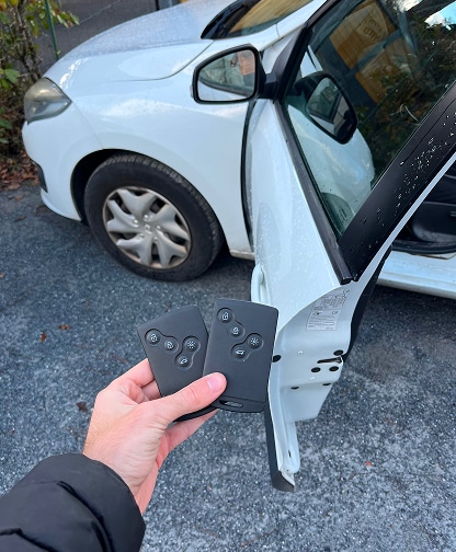 Télécommandes Renault & Voiture Blanche Ouverte Main tenant deux télécommandes Renault noires, près d'une voiture blanche à porte ouverte. Clés de voiture sans clé visibles.